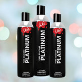 Displayed against a blurred, colorful background are three bottles of Wet Platinum Premium Silicone Lubricant by the brand Wet, ideal for shower sex. This long-lasting silicone lubricant is available in different sizes: 16 fl oz, 9 oz, and 4 oz.
